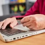 Zwei Männerhande auf der Tastatur eines Laptops. Der Man hat einen roten Pullover an. Eine der beiden Hände hält eine Bankkarte.