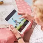 Foto aus der Vogelperspektive: eine ältere Frau hält ein Tablet. Auf dem Tablet ist eine Familie mit kleinen Kindern zu sehen.