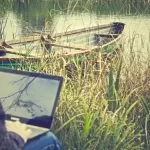 Ein Mann sitzt mit seinem Laptop am See. Hinter dem Bildschirm des Laptops ist ein kleines Holzboot im See.