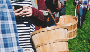 Mehrere Menschen in einer Reihe. Zwei stechen im Bild hervor, sie haben einen Korb am Arm und in der anderen Hand ein Smartphone