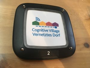 Ein Pager liegt auf einem Tisch. Darauf abgebildet das Logo und Schriftzug von "Cognitive Village - Vernetztes Dorf".