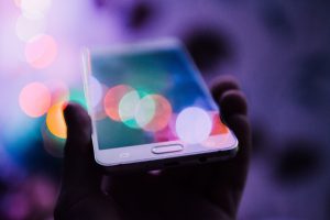 Eine Hand h&auml;lt ein Smartphone hoch. Auf dem Display und im Hintergrund spiegeln sich schillernde Farben, &auml;hnlich wie in einem Kaleidoskop
