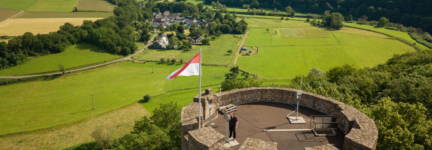 Drohnen-Luftbildaufnahme der Burg Blankenberg in Hennef. Oben auf der Burg ist eine Person,, die an einer der zahlreichen Funkantennen steht.
