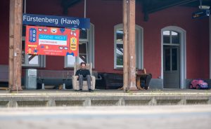 Daniel-Domscheit-Berg sitzt mit einem Laptop auf den Knien auf einem alten Sessel am Gleis des Bahnhofs. Unter dem Bahnhofsschild mit der aufschrift "F&uuml;rstenberg/Havel" ist noch ein gro&szlig;es Schild mit der Aufschrift "Jugend hackt".