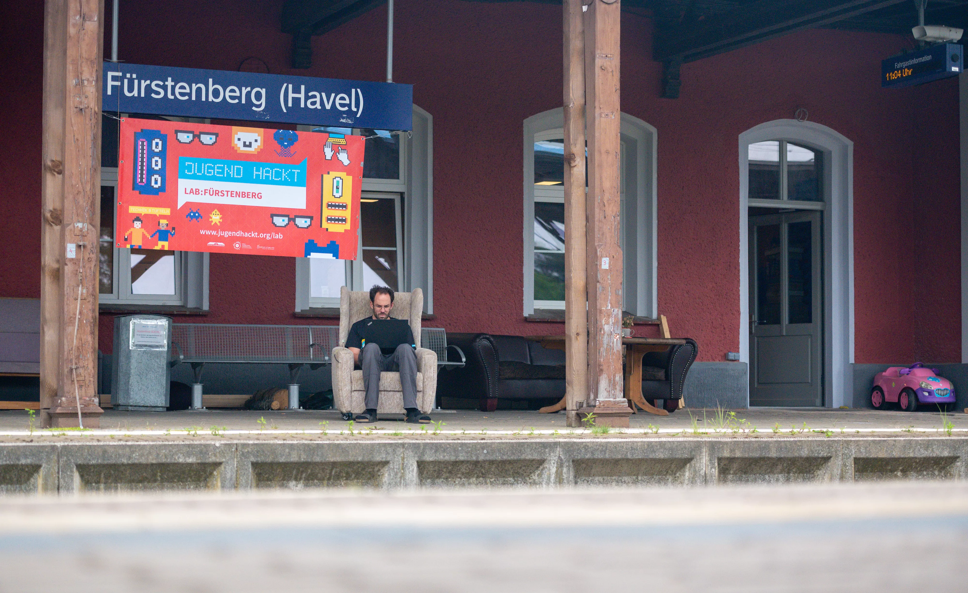 Daniel-Domscheit-Berg sitzt mit einem Laptop auf den Knien auf einem alten Sessel am Gleis des Bahnhofs, dem heutigen Verstehbahnhof. Unter dem Bahnhofsschild mit der aufschrift "Fürstenberg/Havel" ist noch ein großes Schild mit der Aufschrift "Jugend hackt".