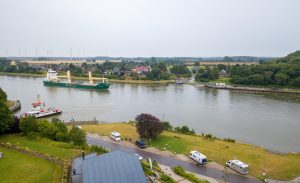 Luftaufnahme aus dem Amt H&uuml;ttener Berge. Im Vordergrund ist ein gr&ouml;&szlig;eres Geb&auml;ude mit ein paar PKWs davor. Im oberen Teil des Bildes ein Fluss mit einem Frachtschiff.