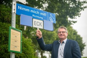 Amtsdirektor Andreas Betz an der Mitfahrbank. Er h&auml;lt die rechte Hand an ein Schild mit der Abk&uuml;rzung ECK f&uuml;r Eckernf&ouml;rde. Dar&uuml;ber ist ein blaues Schild mit der Aufschrift "Nimm mich mit!".