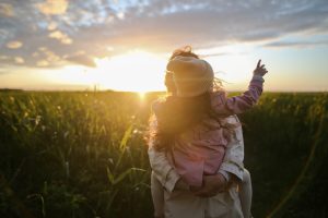 Sonnenuntergang im Hintergrund und ein landwirtschaftlich genutztes Feld im Vordergrund. Ganz vorne ist eine Mustter mit einem Kind auf dem Arm. Man sieht nur den R&uuml;cken des Kindes.