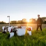 Männlicher Ingenieur mit einem Landwirtschaftsroboter auf einem Feld. Im hintergrund geht die Sonne unter.