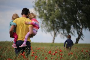 Ein Vater tr&auml;gt links und rechts seine beiden kleinen M&auml;dchen durch ein Feld. Weiter vorne sind zwei B&auml;ume zu sehen. Auf die l&auml;uft sein vorauseilender Sohn zu.